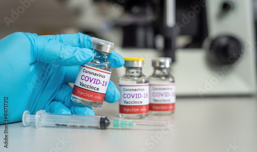 Fototapeta Naklejka Na Ścianę i Meble -  The coronavirus vaccine concept, covid-19, hand held in a blue glove, holding a vial of vaccine placed on a table with a syringe and microscope, inside a medical laboratory.