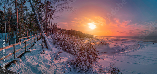 Fototapeta Naklejka Na Ścianę i Meble -  Colorful sunset over snowy sea coast and pine forest with wooden boardwalk path. Beautiful view of Baltic sea coast with snow-covered fir, spruce and pine trees and footpath for relaxing walk.