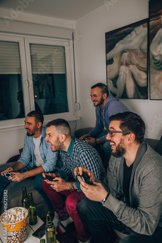 Four happy men playing games and drinking beer while sitting on sofa.