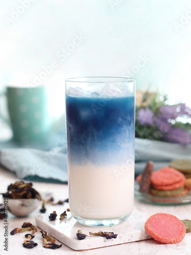 A glass of iced butterfly pea flower milk tea latte with colorful biscuits and its dried flowers on a table, it's a herbal and caffeine free drink