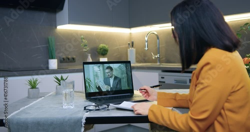 Back view of attractive attantive successful dark-haired woman in orange jacket which sitting at kitchen table and has online video conference with partner photo