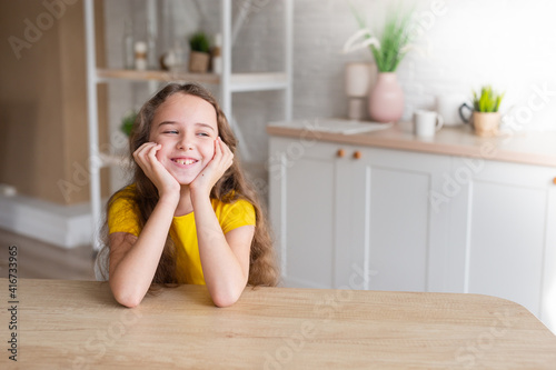 cute school-age girl with curly-haired girls sits at a table in the kitchen, has fun, singing while waiting for food, a delighted child relaxes at the counter, enjoys a happy childhood at home 