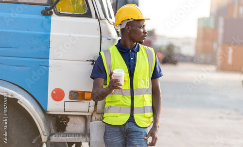 Black worker wearing an engineer hat and a uniform shirt holds a smartphone to the container yard to inspect the logistics import of the country.