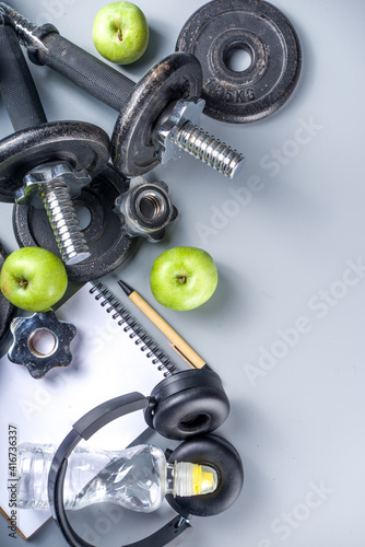 Mens Fitness background with dark Iron dumbbells, sneakers, headphones, gren apples and water bottle. On grey background top view copy space
