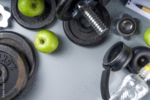 Mens Fitness background with dark Iron dumbbells, sneakers, headphones, gren apples and water bottle. On grey background top view copy space