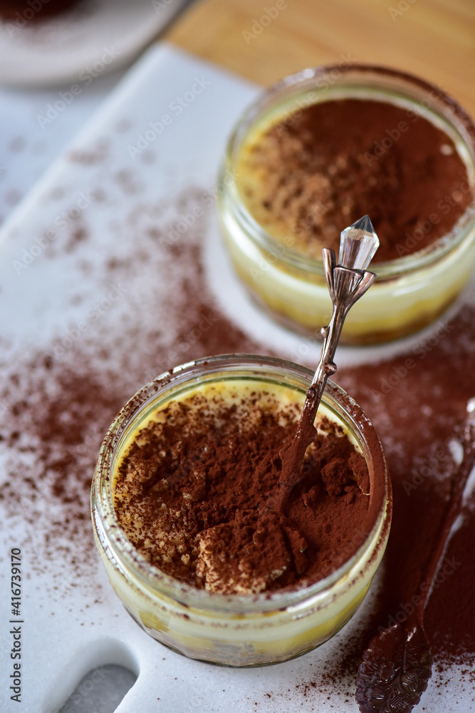 Two jars of cheesecake, sprinkled with cocoa powder, on a white marble board, with a teaspoon. Two jars of cheesecake, sprinkled with cocoa powder, on a white marble board, with a teaspoon.