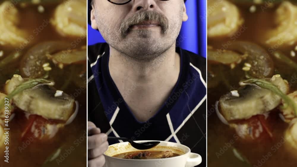 Close-up of a mustachioed man eating tom yam. Thick red soup. Spoon with shrimp. Close-up of tom yam. Blue background.