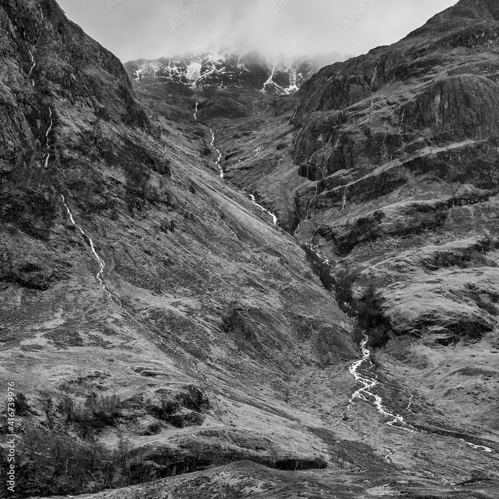 Majestic moody  black and white landscape image of Three Sisters in Glencoe in Scottish Highlands on a wet Winter day wit high water running down mountains