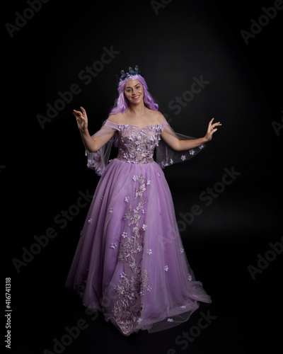 Full length portrait of girl wearing long purple fantasy ball gown with crown and pink hair, standing pose with elegant gestural movements against a studio background.
