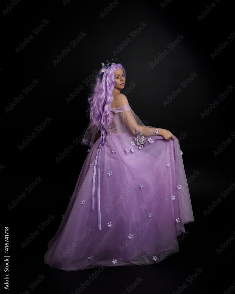 Full length portrait of girl wearing long purple fantasy ball gown with crown and pink hair, standing pose with elegant gestural movements against a studio background. Full length portrait of girl wearing long purple fantasy ball gown with crown and pink hair, standing pose with elegant gestural movements against a studio background.