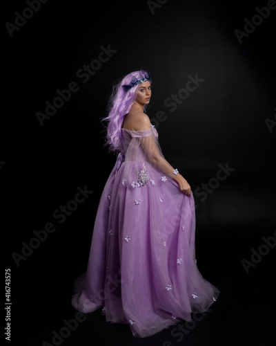 Full length portrait of girl wearing long purple fantasy ball gown with crown and pink hair, standing pose with back to the camera against a studio background.