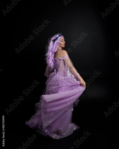 Full length portrait of girl wearing long purple fantasy ball gown with crown and pink hair, standing pose with back to the camera against a studio background.