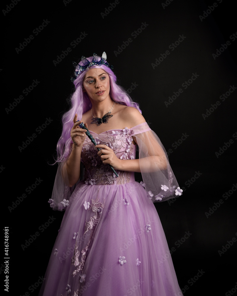 Close up portrait of girl wearing long purple fantasy ball gown with butterfly crown and pink hair, against a  dark studio background.
