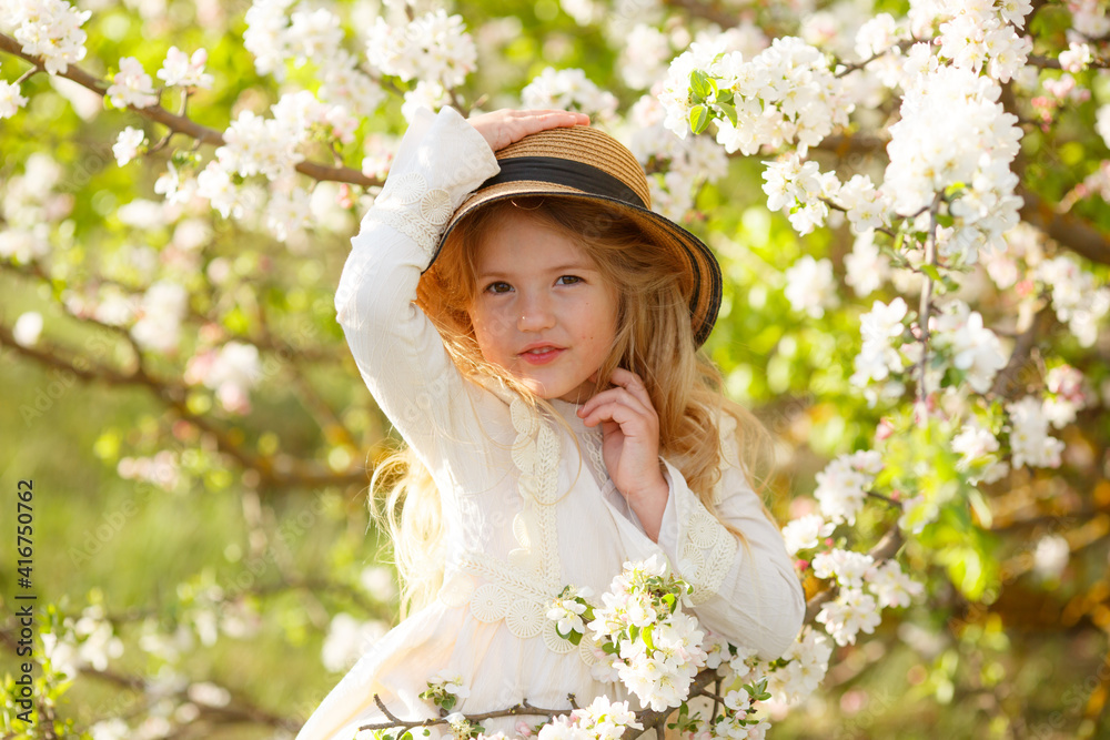 A sweet happy little girl in a white dress in a straw hat in spring near a flowering tree looks at the camera and laughs A sweet happy little girl in a white dress in a straw hat in spring near a flowering tree looks at the camera and laughs