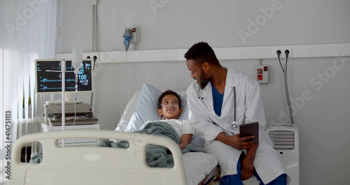 Young african doctor sitting on hospital bed and talking to preteen boy patient