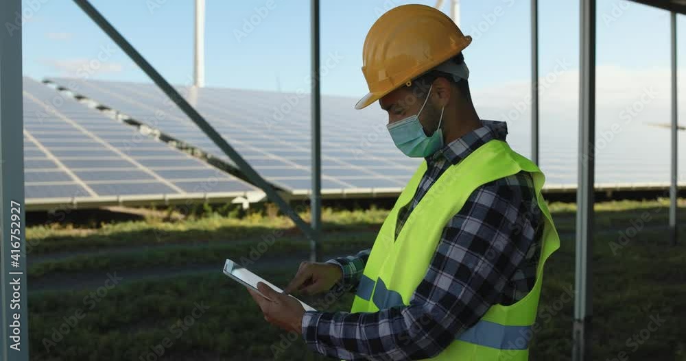 Young man working with digital tablet on solar power station while wearing safety face mask for coroanvirus outbreak Young man working with digital tablet on solar power station while wearing safety face mask for coroanvirus outbreak