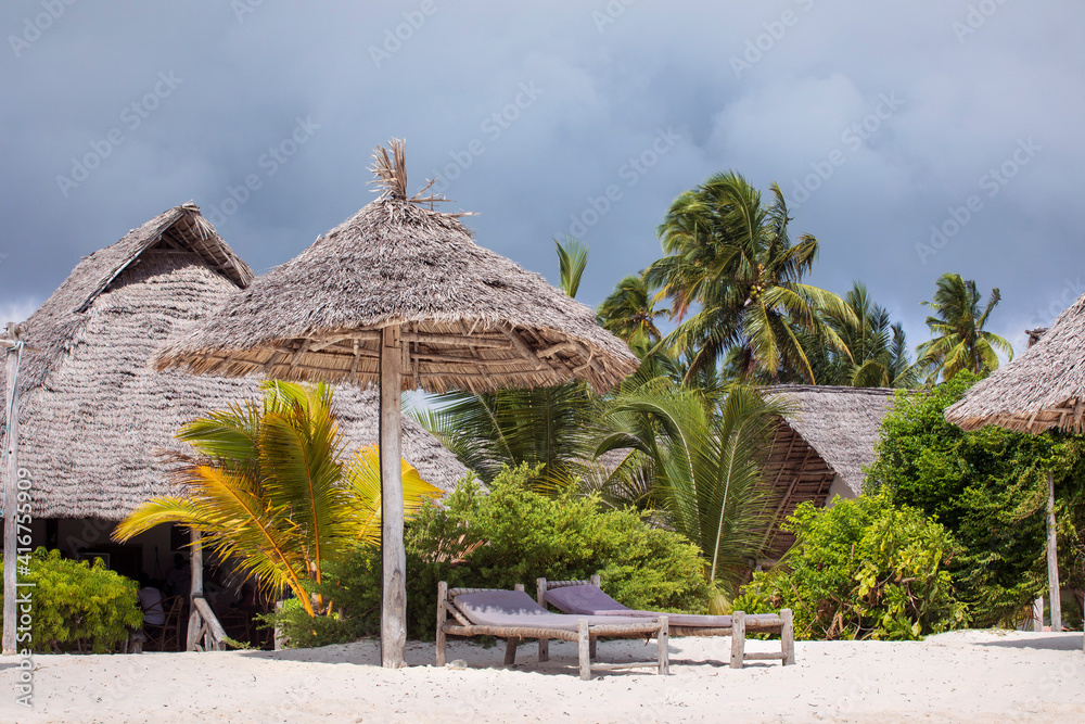 Tourist resort by tropical beach with white sand under palm trees. Sunbeds and straw sun umbrella. Tropical climate dream holidays. 