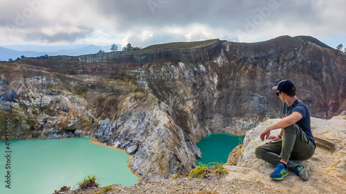 Man sitting at the volcano rim and watching the Kelimutu volcanic crater lakes in Moni, Flores, Indonesia. Man is relaxed and calm, enjoying the view on turquoise lakes. Lakes have different colours photo