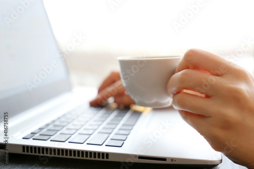 Woman drinks coffee sitting at laptop keyboard on window background. Cup of hot drink in female hand in sunlight, good morning, break during work