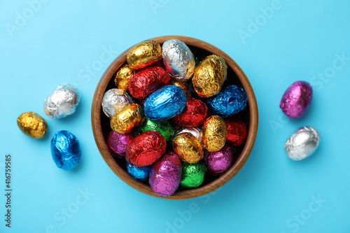 Wooden bowl with chocolate eggs wrapped in colorful foil on light blue background, flat lay