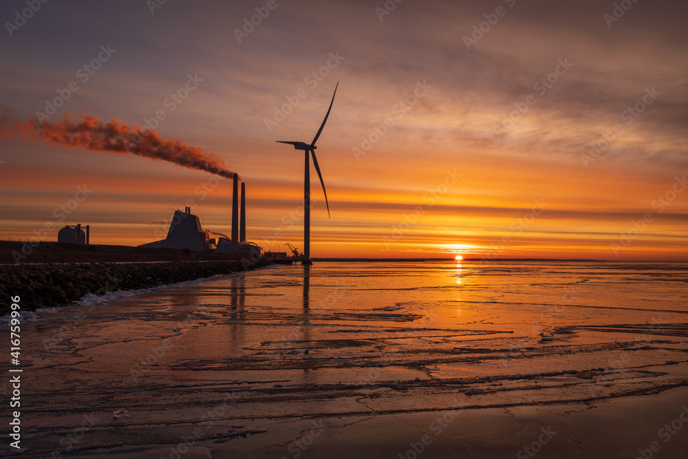 A colourful sunrise at Oresund with a powerplant and a modern Windmill close to the shore. The water is covered with ice reflecting the colours.