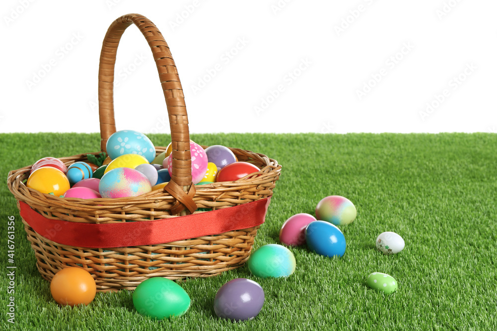 Wicker basket with Easter eggs on green grass against white background. Space for text
