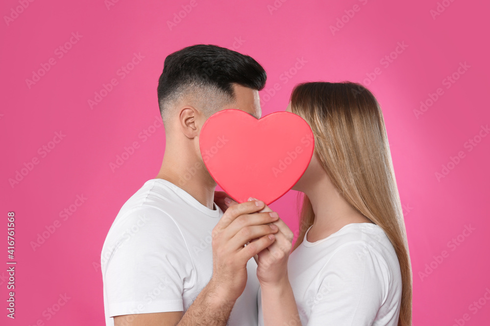 Lovely couple kissing behind decorative heart on pink background. Valentine's day celebration Lovely couple kissing behind decorative heart on pink background. Valentine's day celebration