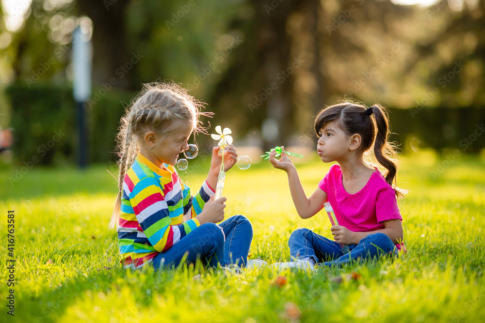 Two little blonde girls and brunette summer sit on the lawn blowing soap bubbles. European and Indian ethnicity children