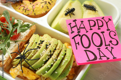 Lunchbox with fake spider, bugs and Happy Fools' Day note on table, closeup