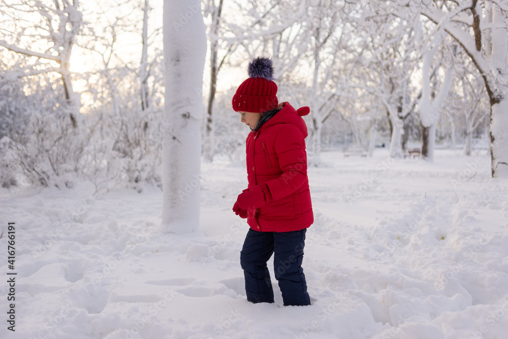 Little Caucasian girl in a red knitted hat and jacket walks in the snow in the park in winter. Winter fun for children and frosty air.