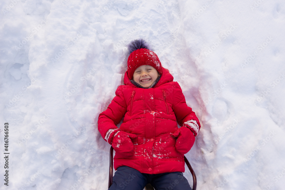 A little girl in a red jacket and a knitted hat lies on the white snow. Children's entertainment in winter. A little girl in a red jacket and a knitted hat lies on the white snow. Children's entertainment in winter.