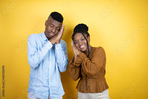 Young african american couple standing over yellow background sleeping tired dreaming and posing with hands together while smiling with closed eyes.