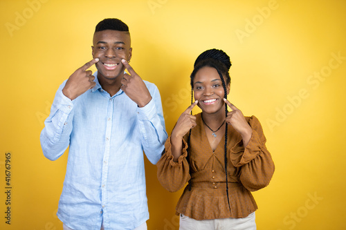 Young african american couple standing over yellow background smiling confident showing and pointing with fingers teeth and mouth