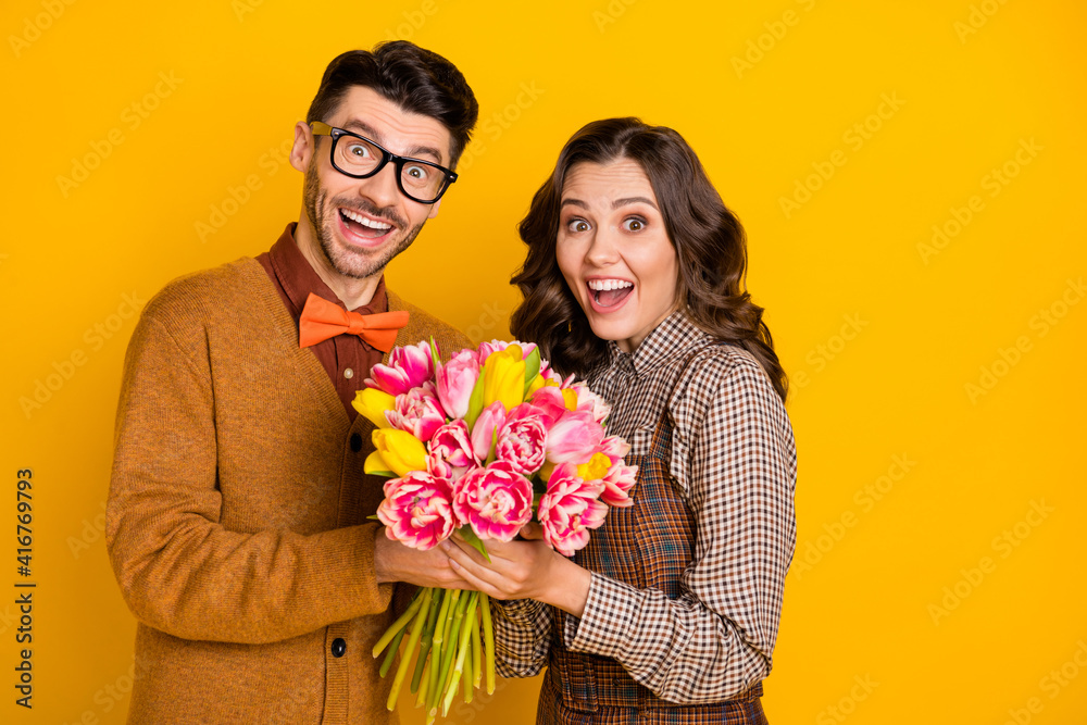 Photo of amazed shocked young man and woman hold hands flowers holiday isolated on bright yellow color background