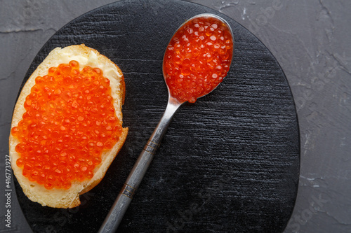 One toast with butter and red caviar near a spoon with caviar on a wooden board on a concrete background. Copy space