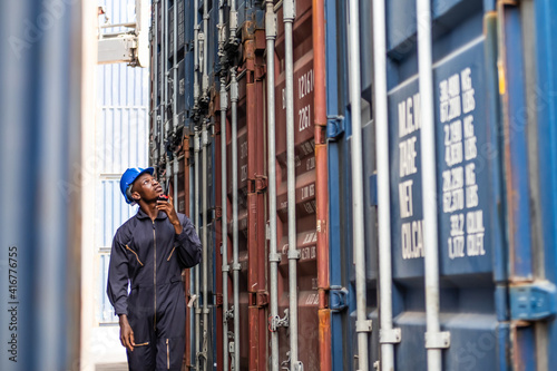 Selective focus at face of Black African logistic worker wearing safety equipment, check and inspection the condition of container while talking and communicate with his team by  radio device. © BritCats Studio