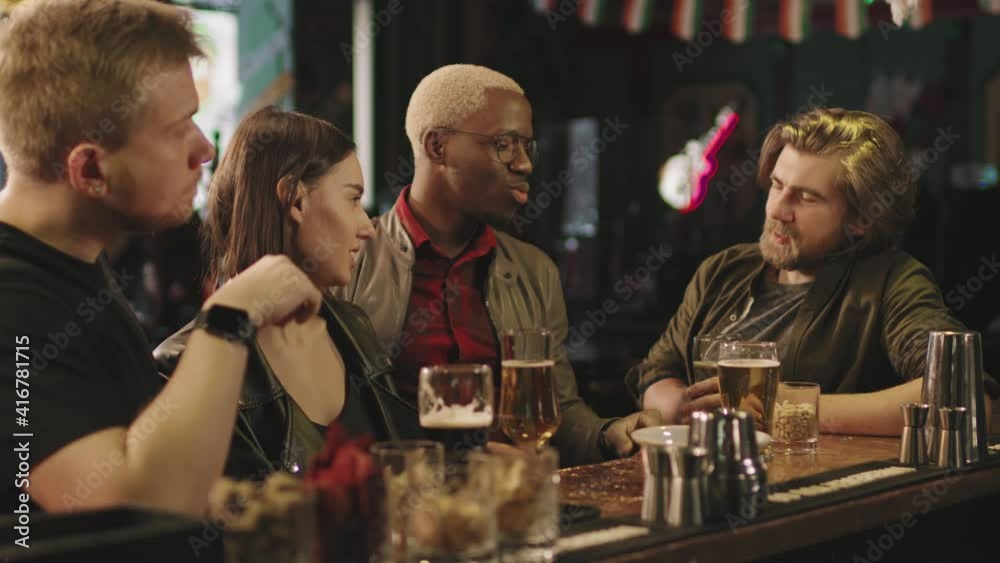 Waist-up footage of happy young group of multi-ethnic friends having fun together sitting at bar drinking beer and chatting