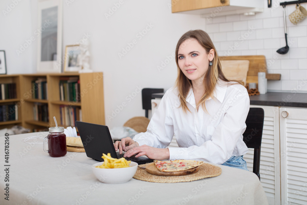 young woman working with laptop and eating fast food in the kitchen. Unhealthy Lifestyle woman has pizza for lunch. home office. young woman working with laptop and eating fast food in the kitchen. Unhealthy Lifestyle woman has pizza for lunch. home office.