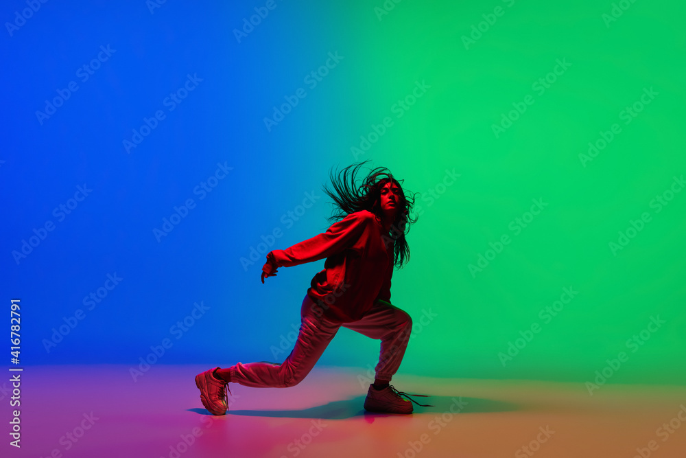Moving. Stylish sportive girl dancing hip-hop in stylish clothes on colorful background at dance hall in neon light. Youth culture, movement, style and fashion, action. Fashionable bright portrait.