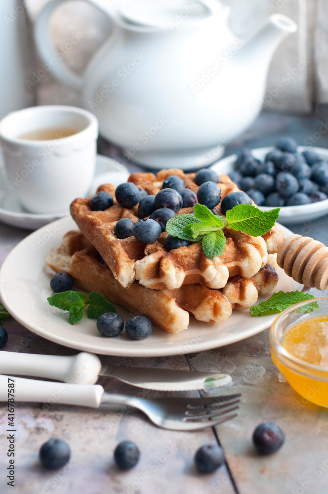Belgian waffles with blueberries, honey and mint on a white plate. Breakfast with a cup of tea with a white teapot and sweet waffles. Knife and fork with white handles. Belgian waffles with blueberries, honey and mint on a white plate. Breakfast with a cup of tea with a white teapot and sweet waffles. Knife and fork with white handles.