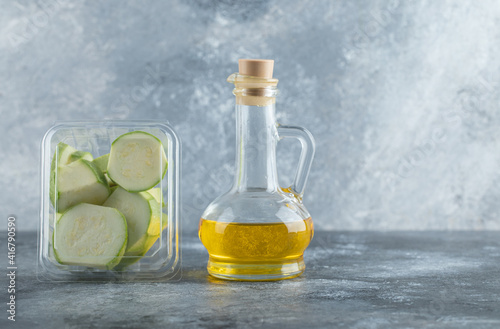Plastic container full with zucchini pieces and bottle of oil over grey background