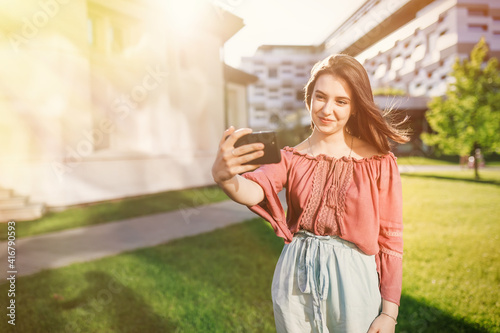 Free Download Beautiful girl with a phone in her hands. The girl looks at herself in the phone camera and takes a selfie. She smiles and enjoys the warm day. - GFXLoad