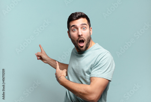 young handsome adult man feeling shocked and surprised, pointing to copy space on the side with amazed, open-mouthed look photo