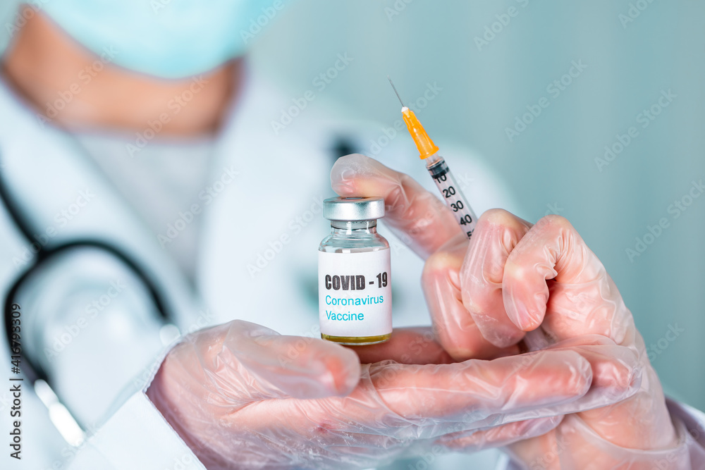 Doctor or nurse in uniform and gloves wearing face mask protective in lab holding medicine vial vaccine bottle with COVID-19 Coronovirus vaccine label Doctor or nurse in uniform and gloves wearing face mask protective in lab holding medicine vial vaccine bottle with COVID-19 Coronovirus vaccine label