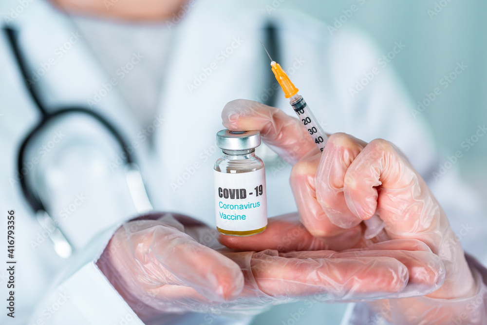 Doctor or nurse in uniform and gloves wearing face mask protective in lab holding medicine vial vaccine bottle with COVID-19 Coronovirus vaccine label Doctor or nurse in uniform and gloves wearing face mask protective in lab holding medicine vial vaccine bottle with COVID-19 Coronovirus vaccine label