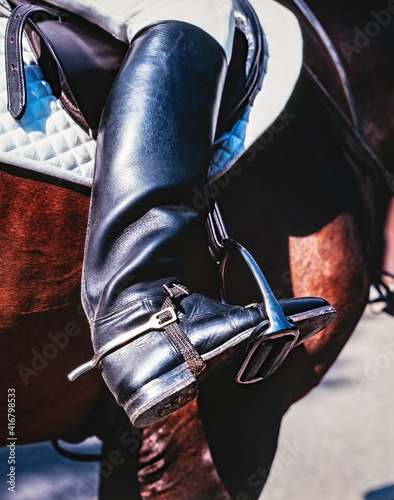 A rider's foot on a black horse close up. A woman's booted foot standing in a black stirrup of horse saddle.
