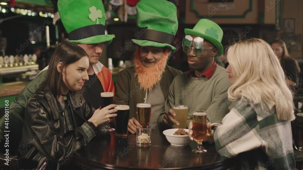 Medium shot of cheerful young group of multi-ethnic friends celebrating St Patricks Day in pub drinking beer. Men wearing Irish costumes and laughing, while women chatting