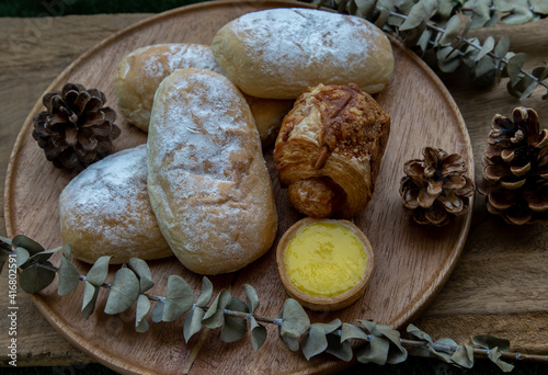Homemade sweet cream soft buns, Baked croissant sausage and Lemon tart with Dried flowers on round wooden plate over rustic wooden floor. Bakery and dessert concept, Selective focus.