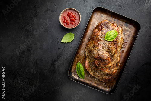 Baked pork meat in a baking pan on dark background, top view