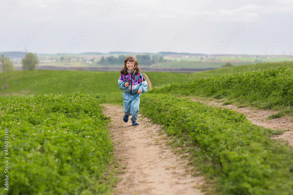 A little girl runs along a field dirt road. She laughs, her hair tousled by the wind. Lots of green grass on background